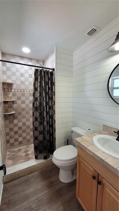 Bathroom featuring white shiplap walls, wood-finish flooring, a tiled vanity countertop with an integrated basin, and a walk-in shower with built-in shelving and multi-toned wall tile