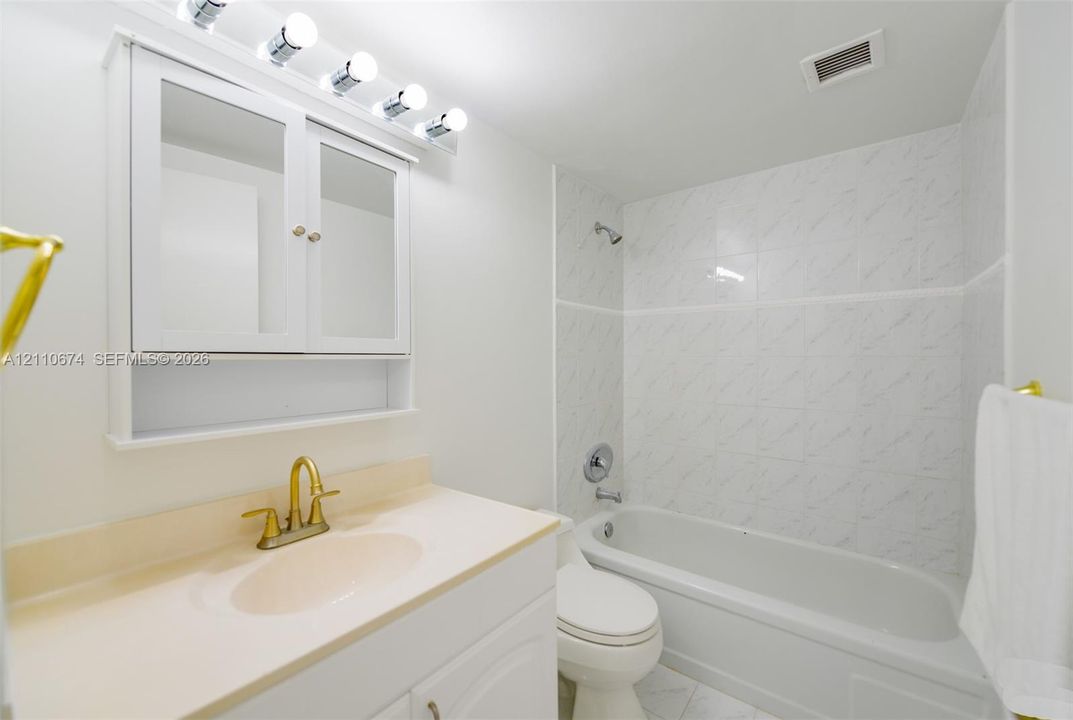 Bathroom featuring a vanity with integrated countertop and brass-finish faucet, white wall-mounted medicine cabinet with integrated lighting, and a shower-tub combination with patterned tile surround