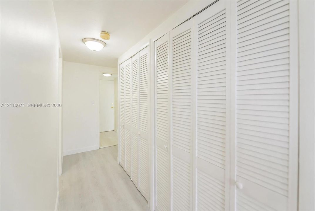 Full-length hallway featuring white louvered bifold closet doors, light-toned wood-finish flooring, and a flush-mount ceiling light fixture