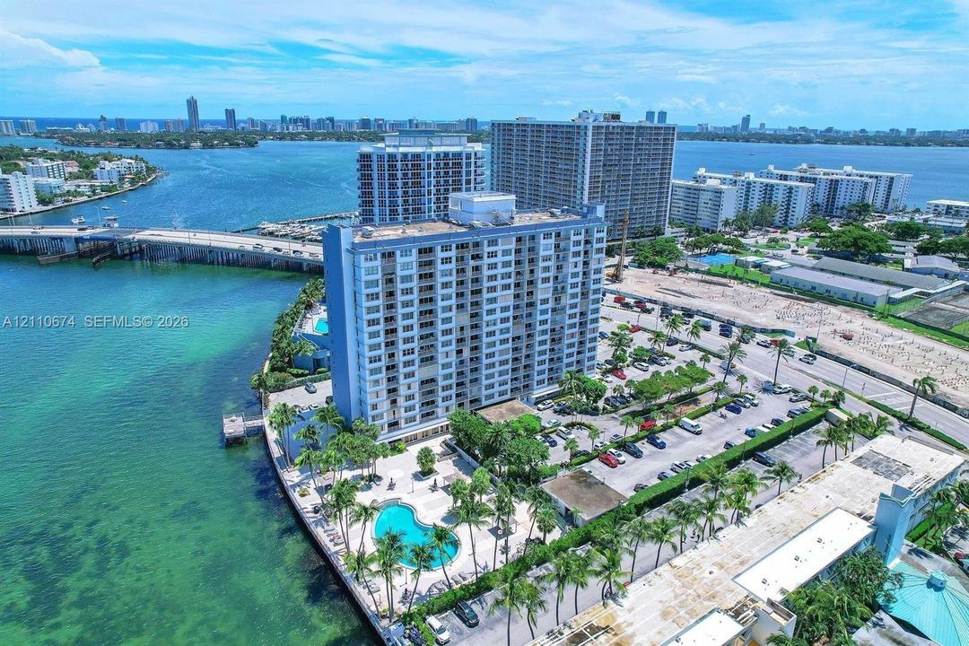 High-rise condominium building featuring a waterfront swimming pool, landscaped palm trees, paved parking areas, and expansive water frontage