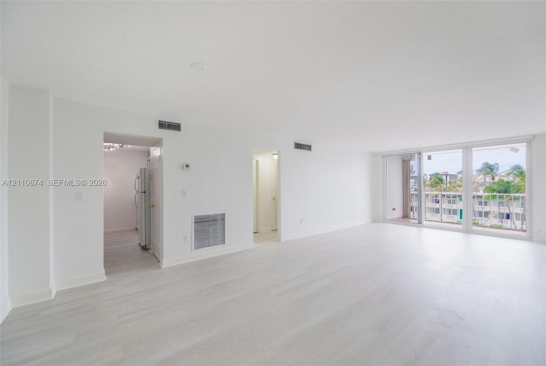 Spacious open-concept interior featuring wood-finish flooring, white walls, and floor-to-ceiling glass sliding doors leading to an exterior balcony