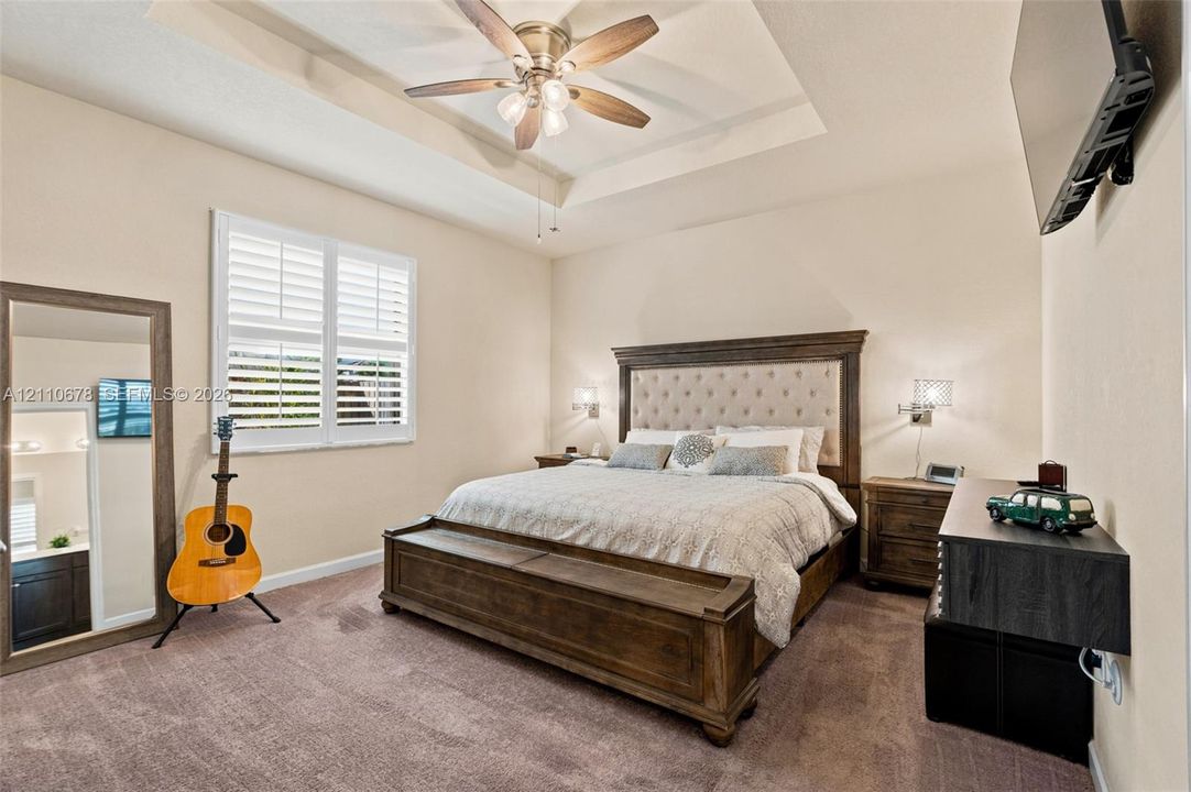 Tray ceiling with a central multi-blade ceiling fan, wall-mounted shuttered windows, and neutral carpeting