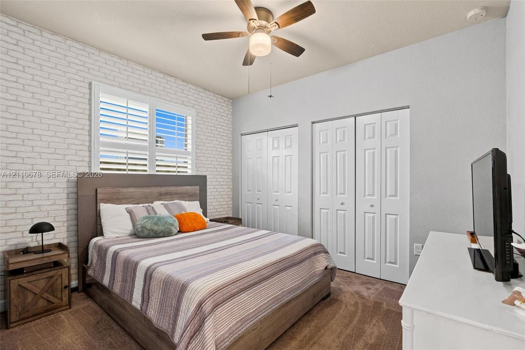 Room featuring an accent wall with white brick-style masonry, two sets of white bi-fold closet doors, wall-to-wall carpeting, and a ceiling fan with integrated lighting