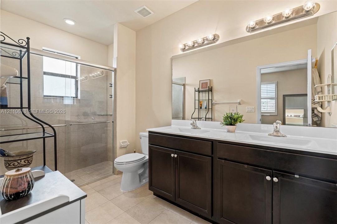 Dark wood vanity featuring dual integrated sinks, white countertop, full-width mirror, and two multi-bulb fixtures, accompanied by a glass-enclosed shower with tile surround and neutral-tone floor tiling