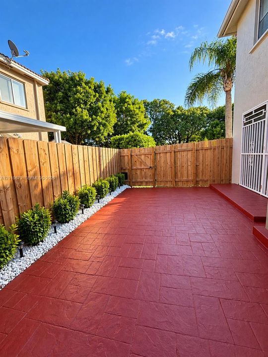 Stamped concrete patio featuring a perimeter of white decorative stones, integrated shrubbery, a wood privacy fence, and a metal security door
