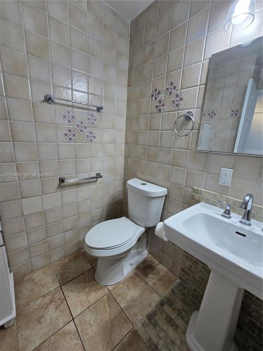 Full-height neutral wall tiling featuring decorative inlay accents, porcelain floor tile, pedestal sink with polished chrome fixtures, and wall-mounted metal hardware