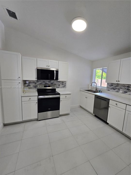 Kitchen featuring white shaker-style cabinetry, stainless steel appliances, patterned tile backsplash, solid-surface countertops, and large-format tile flooring