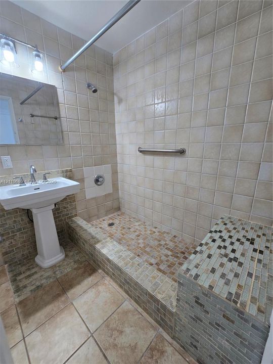 White pedestal sink with chrome widespread faucet, tiled walk-in shower featuring an integrated bench seat, mosaic stone-finish shower floor, and stainless steel grab bar