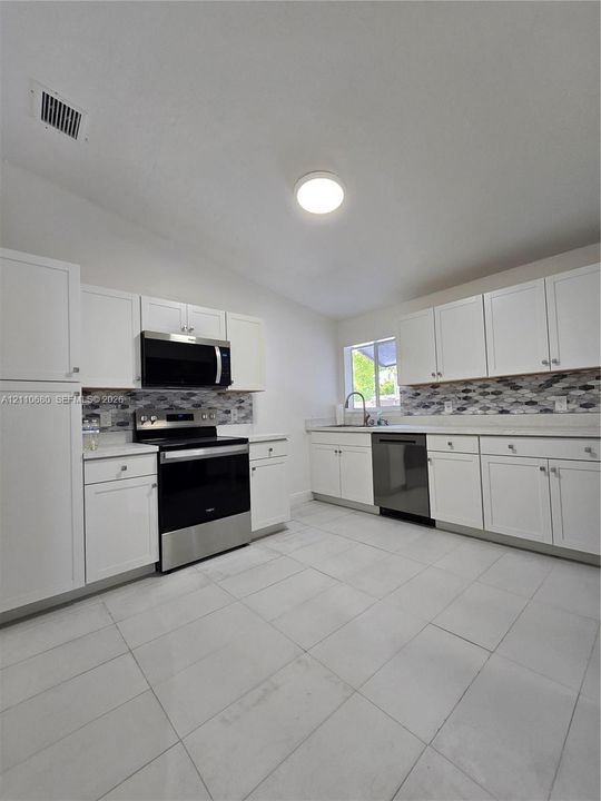 Kitchen featuring white shaker-style cabinetry, stainless steel appliances, patterned mosaic tile backsplash, solid-surface countertops, and light-toned tile flooring