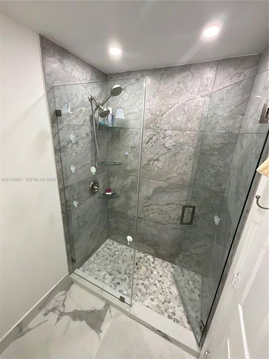 Walk-in shower featuring large-format stone-finish wall tile, pebble-tile flooring, frameless glass enclosure, and built-in glass corner shelving