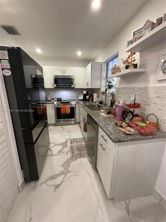 Kitchen featuring white shaker-style cabinetry, stone-finish countertops, stainless steel appliances, stacked-tile backsplash, and large-format porcelain tile flooring