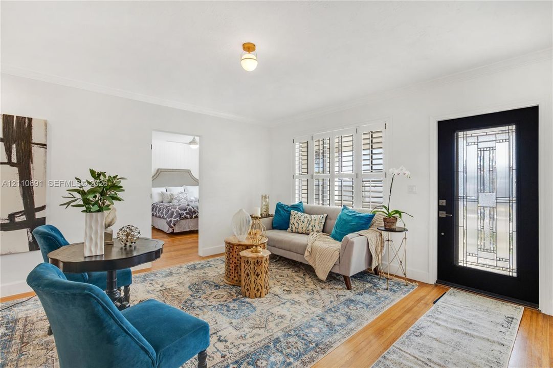 Wood-finish flooring, white walls, multi-pane casement windows with plantation shutters, globe-style ceiling lighting, and a black front door featuring geometric patterned glass inlay