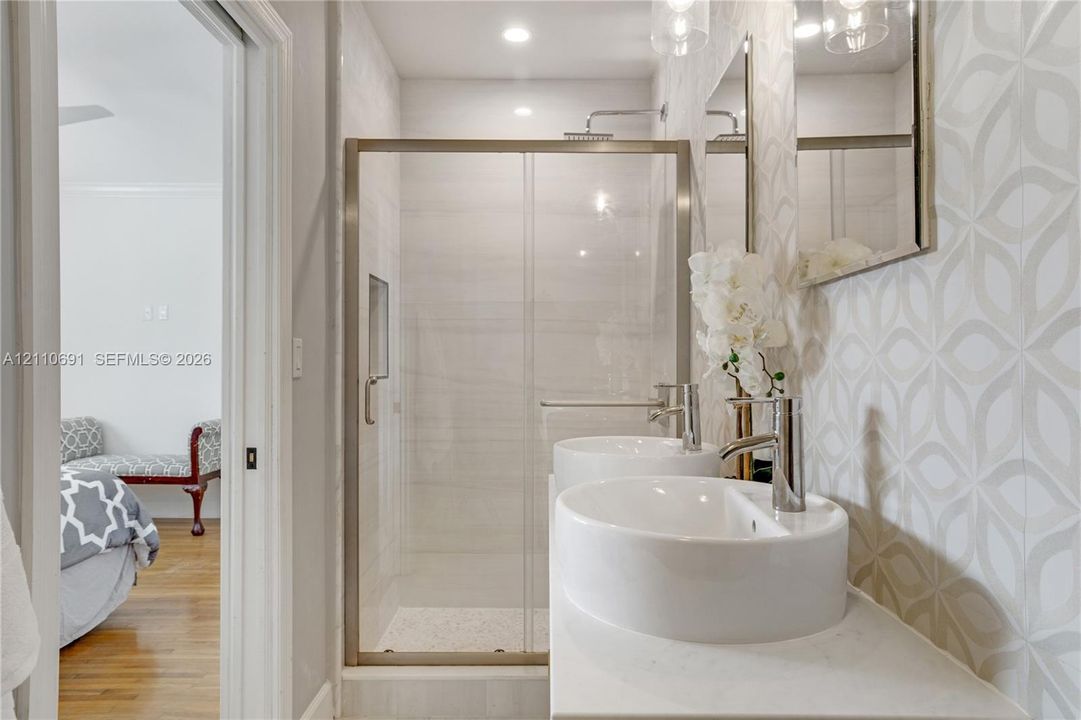 Dual vessel sinks with polished chrome faucets, quartz-surfaced vanity, glass-enclosed shower with recessed shelving, and decorative geometric wall tile