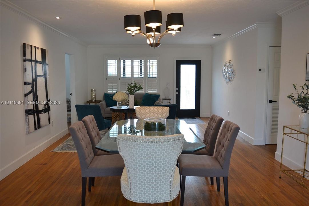 Wood-finish flooring, multi-light chandelier with black shades, white plantation shutters, and a high-contrast black exterior door frame