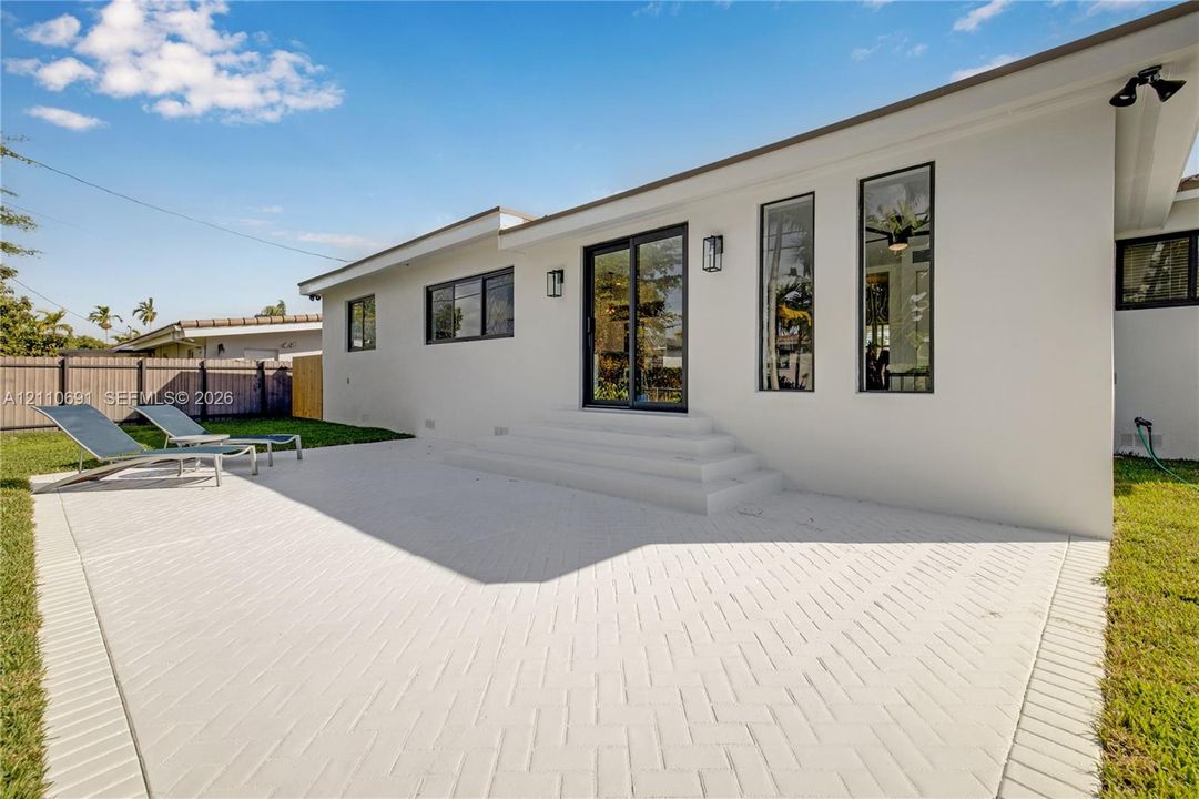 Rear exterior featuring a herringbone-patterned paved patio, white stucco siding, sliding glass doors, vertical rectangular windows, and exterior wall-mounted lighting