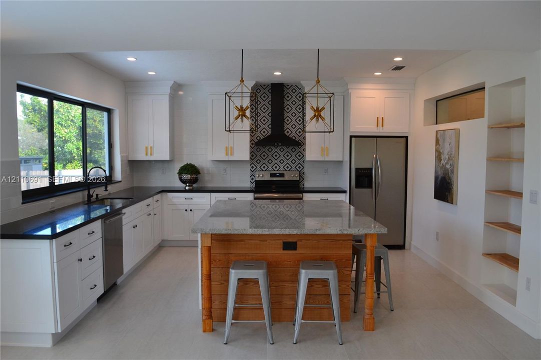 Kitchen featuring white shaker cabinetry, stone-topped central island with wood paneling, geometric tile backsplash, recessed lighting, and built-in shelving
