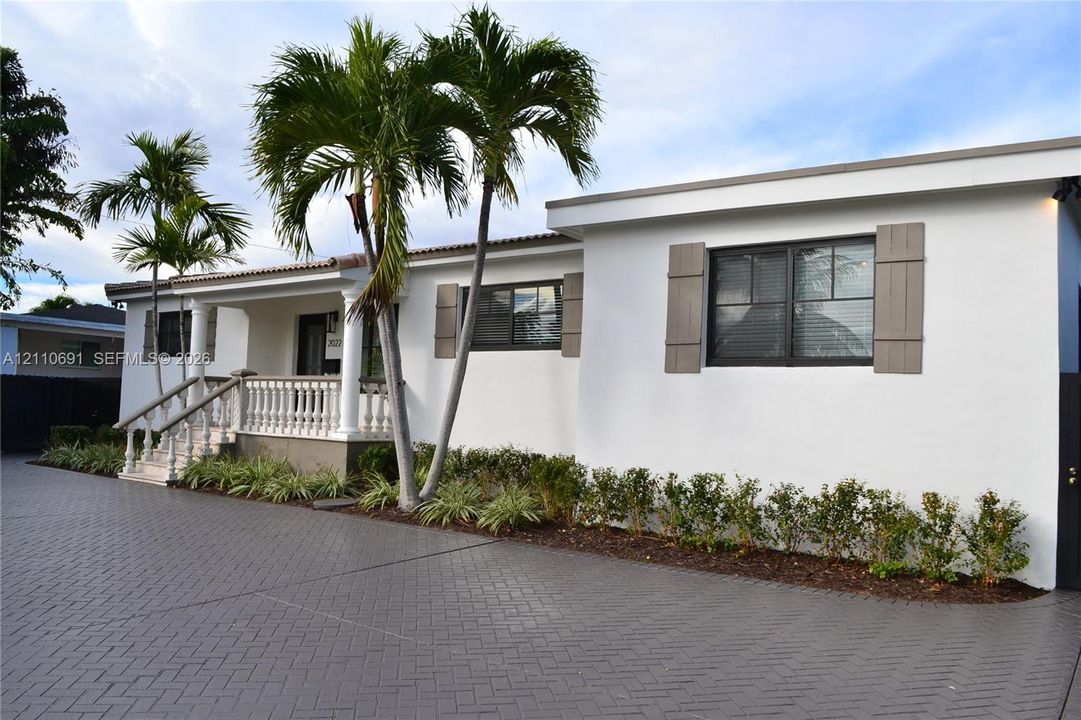 Stucco exterior featuring a covered front porch with a concrete balustrade, herringbone-patterned paved driveway, decorative shutters, and mature palm landscaping