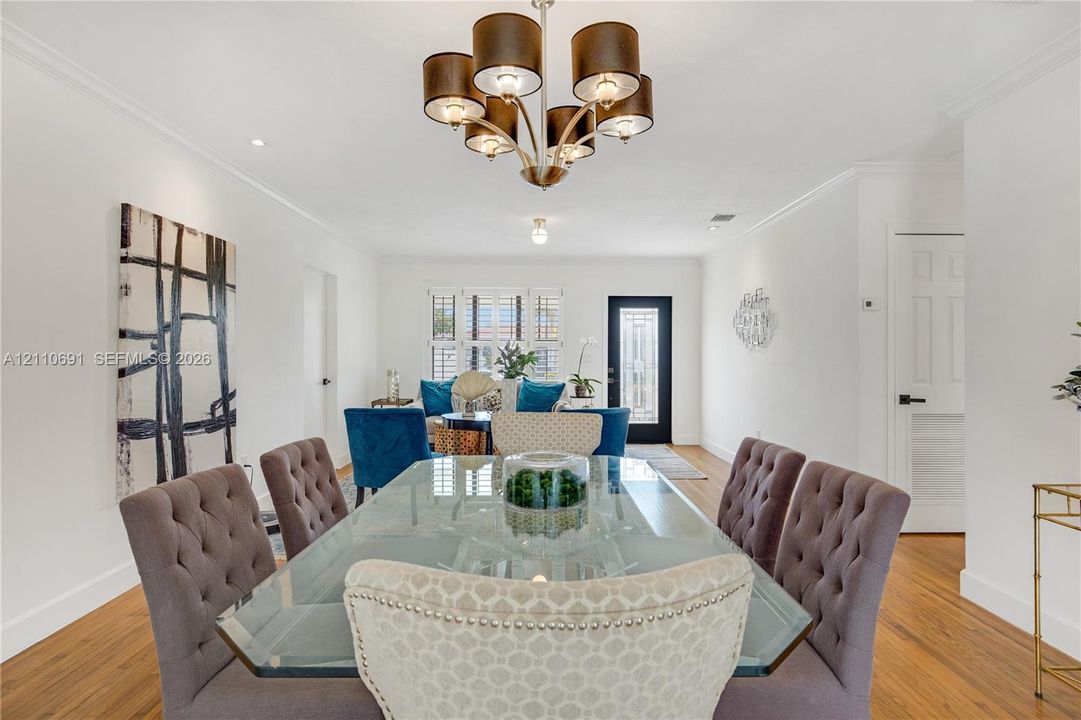 Dining space featuring wood-finish flooring, multi-arm drum shade chandelier, crown molding, plantation shutters, and glass-paneled entry door