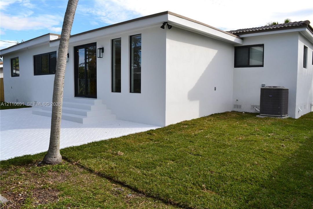 Stucco exterior featuring a white paved patio, tiered concrete steps, dark-framed glass doors, and dark-framed windows