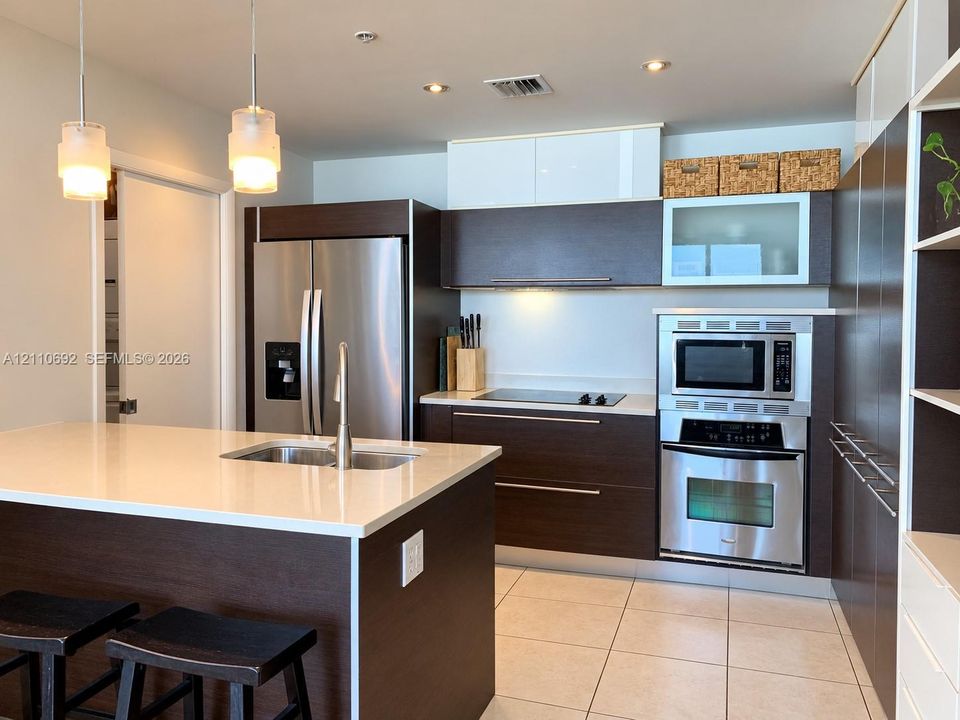 Kitchen featuring dark-finish cabinetry, stone-surface countertops, stainless steel appliances, a central island with a double-basin sink, and recessed ceiling lighting