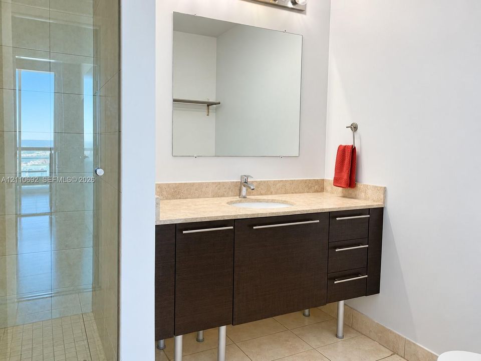 Floating vanity with dark-finish cabinetry, metal legs, stone-look countertop, and integrated sink basin, paired with a frameless mirror and a walk-in shower featuring glass doors and tiled surround