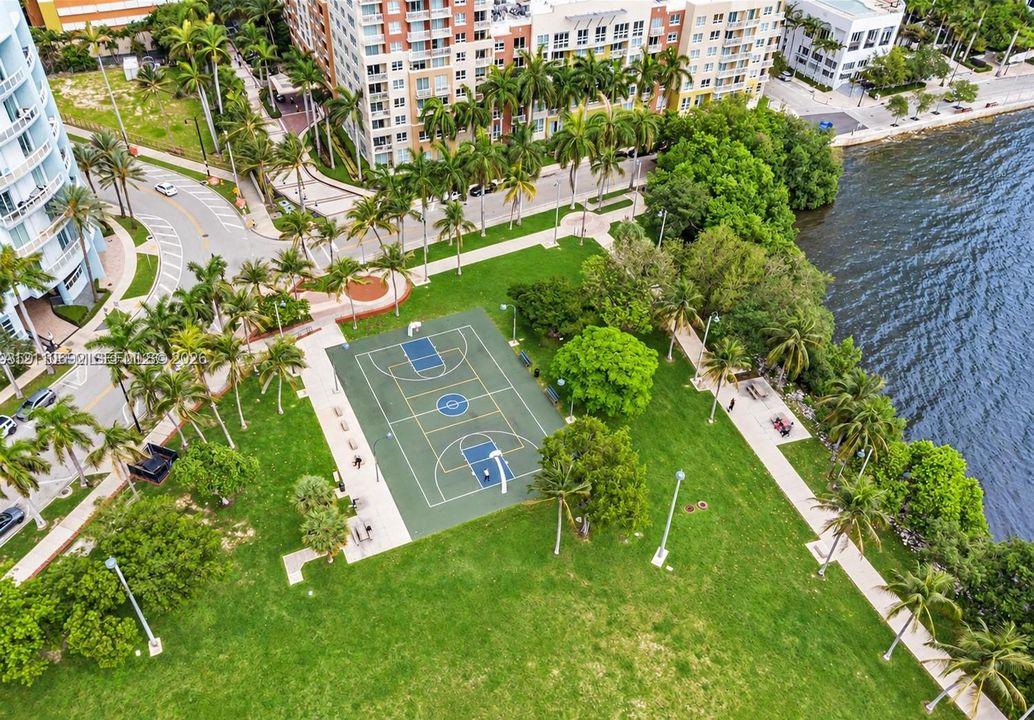Waterfront outdoor space featuring a paved basketball court, manicured lawn, established palm trees, and pedestrian walkways