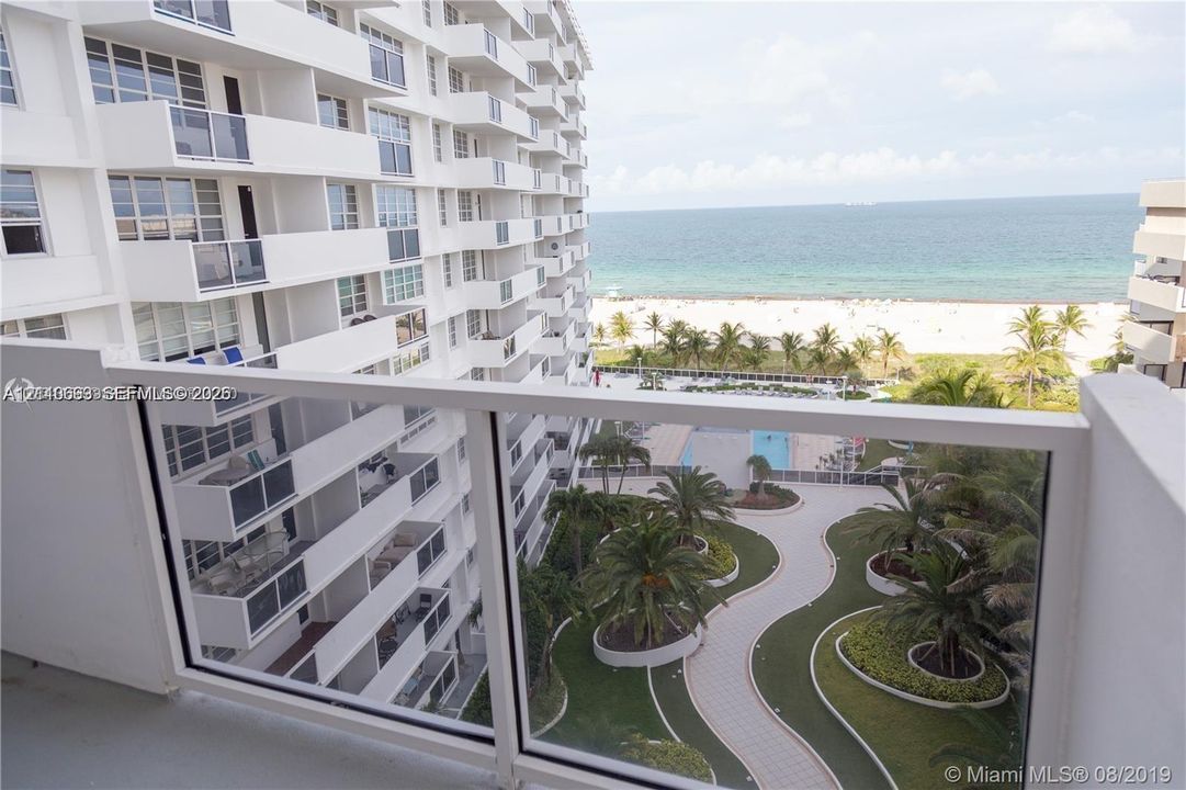 Private balcony with glass panel railing, overlooking a landscaped courtyard, swimming pool, and sandy beach shoreline