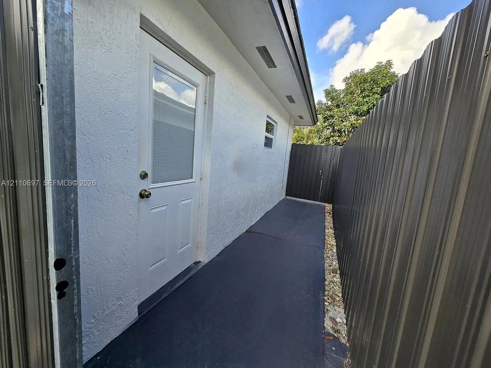 Exterior side yard featuring a smooth stucco finish, a half-glass entry door, a paved walkway, and corrugated metal perimeter fencing