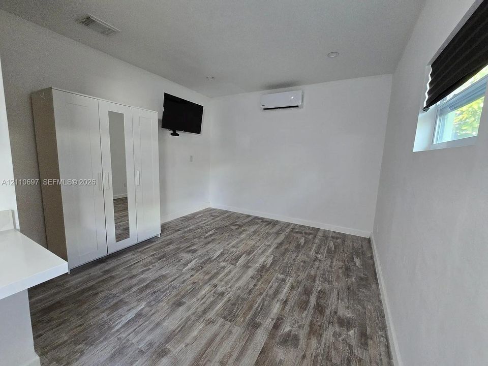 Wood-finish flooring, white walls, recessed ceiling lighting, a wall-mounted climate control unit, and a window with a fitted shade