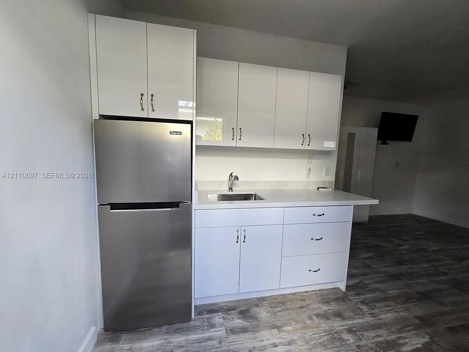 Kitchenette featuring white slab-panel cabinetry, light-toned stone-finish countertops, an undermount stainless steel sink with a high-arch faucet, a stainless steel refrigerator, and wood-finish flooring
