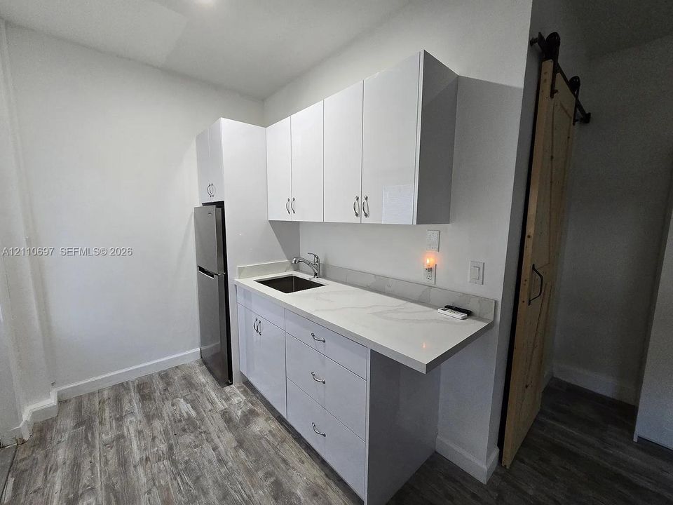 Kitchenette featuring high-gloss white cabinetry, white stone-finish countertops with integrated backsplash, stainless steel sink, and wood-finish flooring with a sliding barn-style door