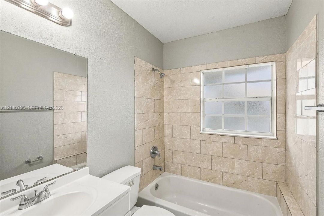 Bathroom featuring a white vanity with an integrated sink, polished chrome faucet, full-width mirror, light fixture, and a tile-surround tub with a frosted window
