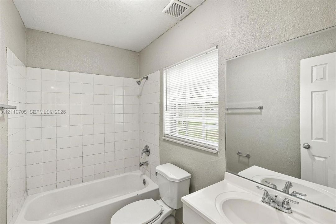 Bathroom featuring a white bathtub with a tiled surround, a built-in vanity with a sink, a wall-mounted mirror, and a window with blinds