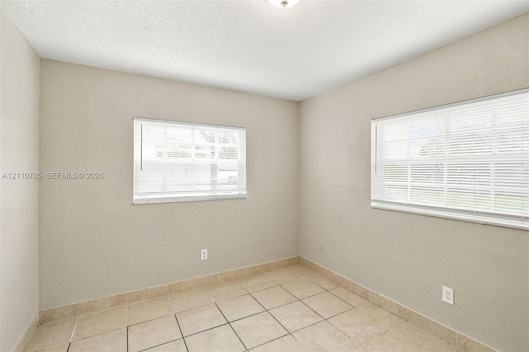 Neutral-toned interior space featuring light-colored tile flooring, smooth wall finishes, two windows with horizontal blinds, and an overhead flush-mount light fixture