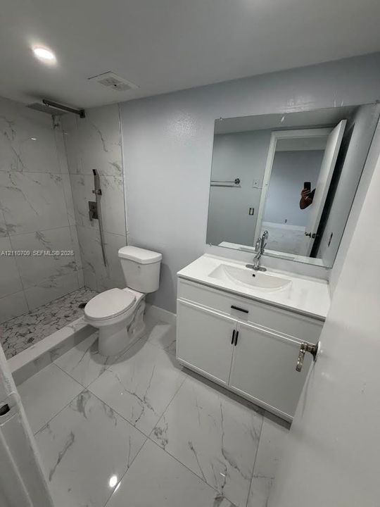 Bathroom featuring large-format tile flooring, a walk-in shower with stone-patterned wall tile, a vanity with a white countertop and integrated sink, and a full-width wall mirror