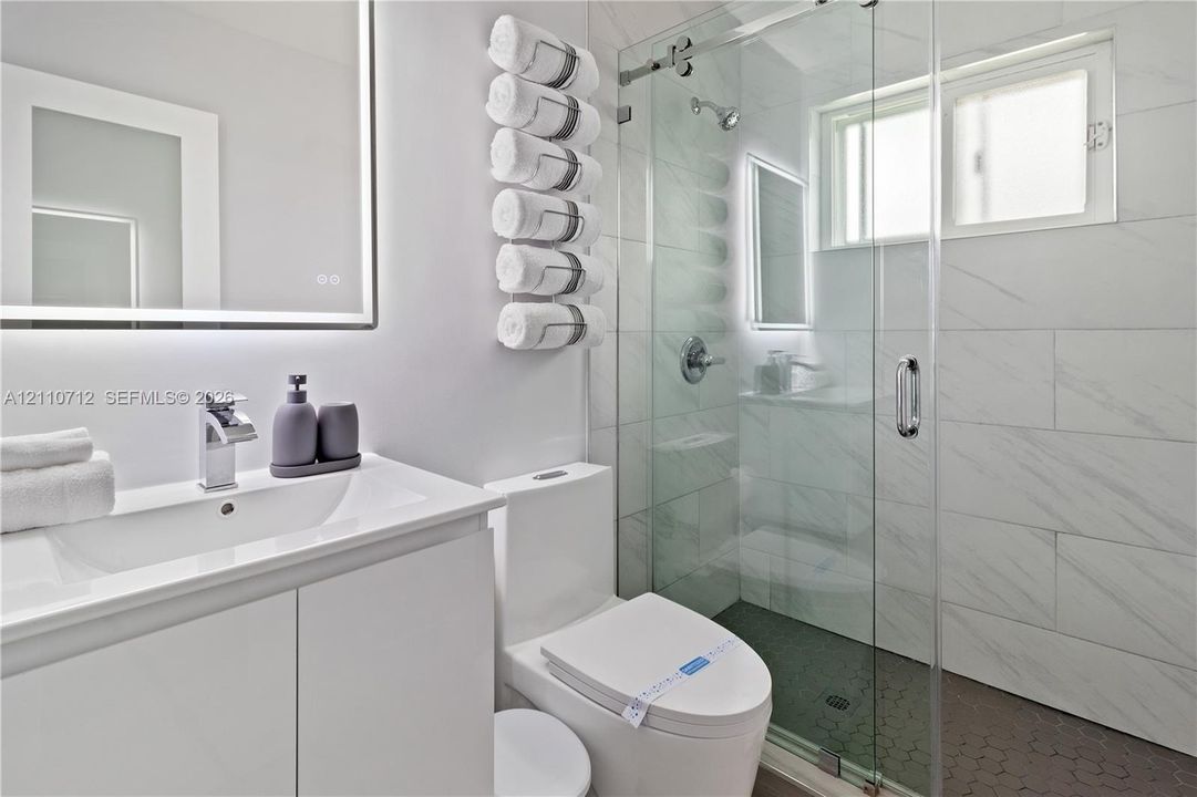 Bathroom featuring a white vanity with integrated basin, LED-backlit mirror, frameless glass shower enclosure, large-format tiled shower walls, and hexagonal floor tiling