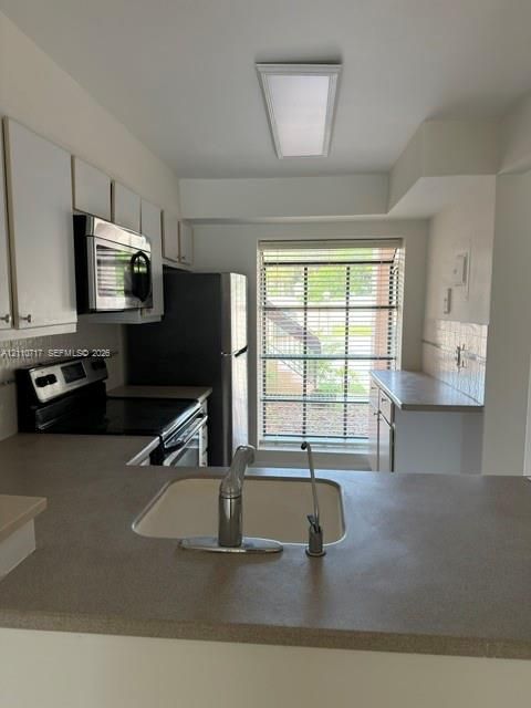 Kitchen featuring flat-panel white cabinetry, solid-surface countertops, integrated stainless steel appliances, and a rectangular window with horizontal blinds