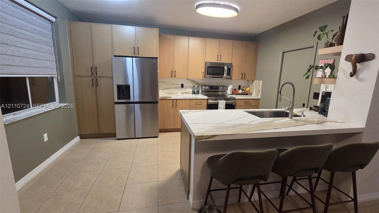 Kitchen featuring wood cabinetry, stone-look backsplash, built-in stainless steel appliances, floating shelves, and tile flooring