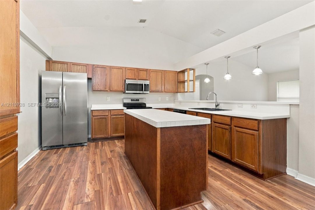 Kitchen featuring wood-finish cabinetry, a central island with tiled countertop, stainless steel refrigerator, microwave, and three pendant lights
