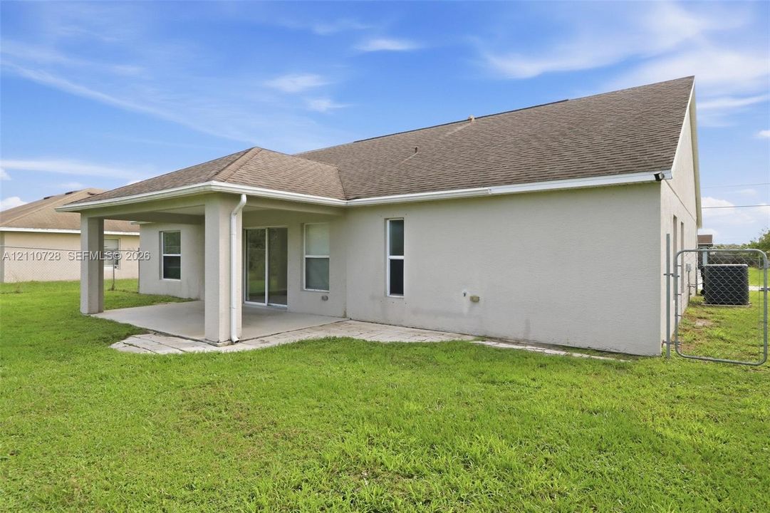 Rear exterior featuring a covered concrete patio, stucco siding, a multi-gabled asphalt shingle roof, and a chain-link fenced perimeter
