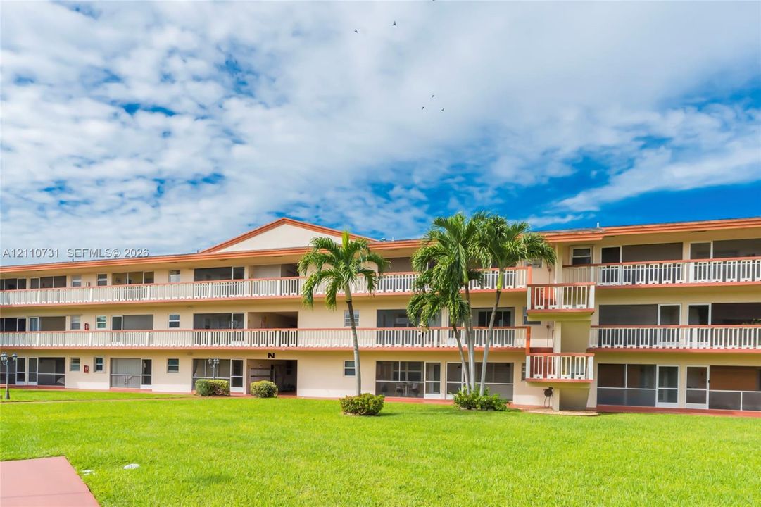 Three-story multi-family exterior featuring stucco siding, wrap-around balconies with white railings, screened-in exterior entryways, and mature palm trees set against a manicured lawn