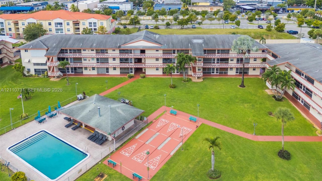 Multi-story residential complex featuring a swimming pool, covered outdoor pavilion, shuffleboard courts, and expansive manicured green space