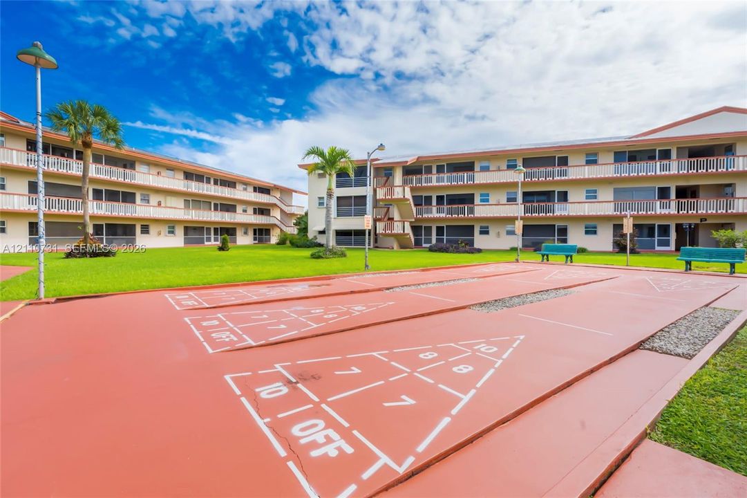 Outdoor shuffleboard court featuring a paved surface, multi-story buildings with external balconies, and landscaped lawn areas