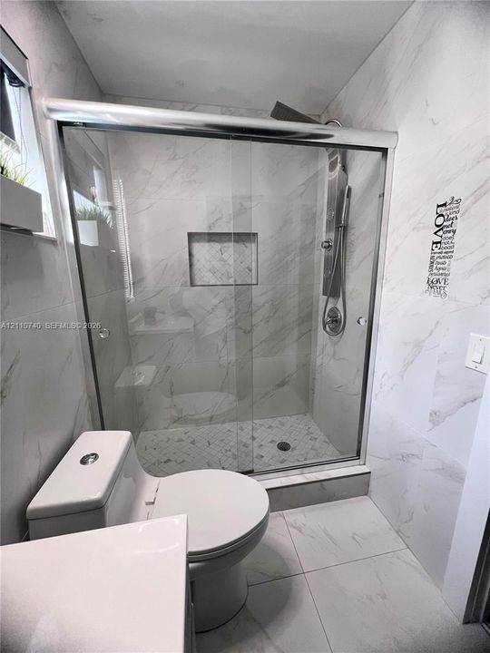 Walk-in shower featuring glass sliding doors, built-in recessed shelving, wall-mounted rain shower head, and floor-to-ceiling stone-finish tile surround