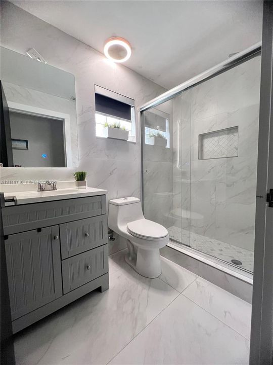 Bathroom featuring grey cabinetry with silver-tone hardware, stone-finish tiling, a glass-enclosed shower with a built-in recessed niche, and a contemporary flush-mount light fixture
