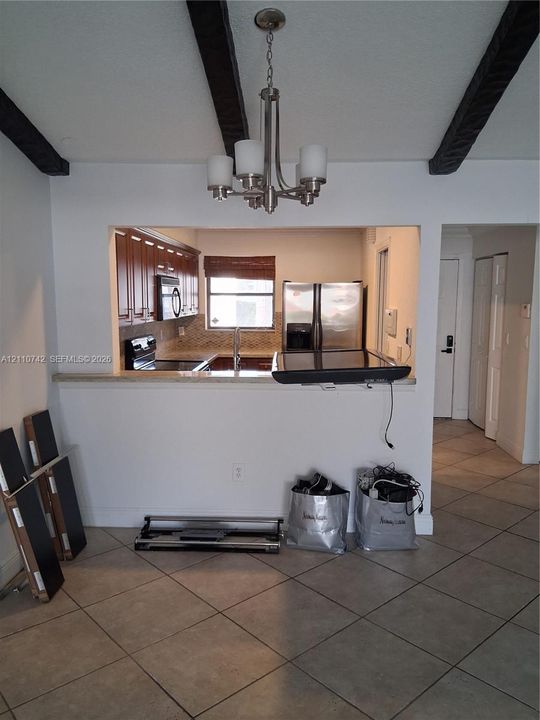 Open-concept interior featuring exposed dark wood-finish ceiling beams, a brushed nickel five-light chandelier, ceramic tile flooring, and a pass-through opening with a stone-finish countertop