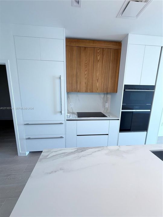 Custom cabinetry featuring contrasting wood-finish uppers and matte white base units, integrated refrigerator, built-in dual oven, stone-surface backsplash and countertops, and large central island