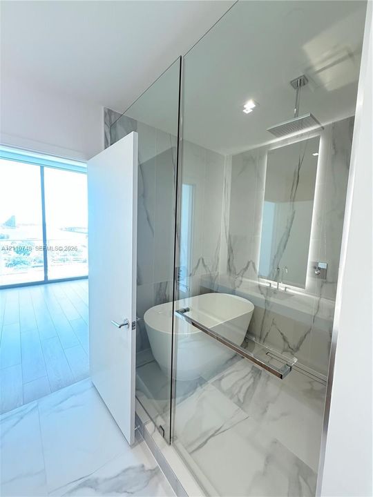 Freestanding soaking tub enclosed in a glass-partitioned wet room featuring floor-to-ceiling stone-look tiling, a ceiling-mounted rainfall showerhead, and a wall-mounted faucet