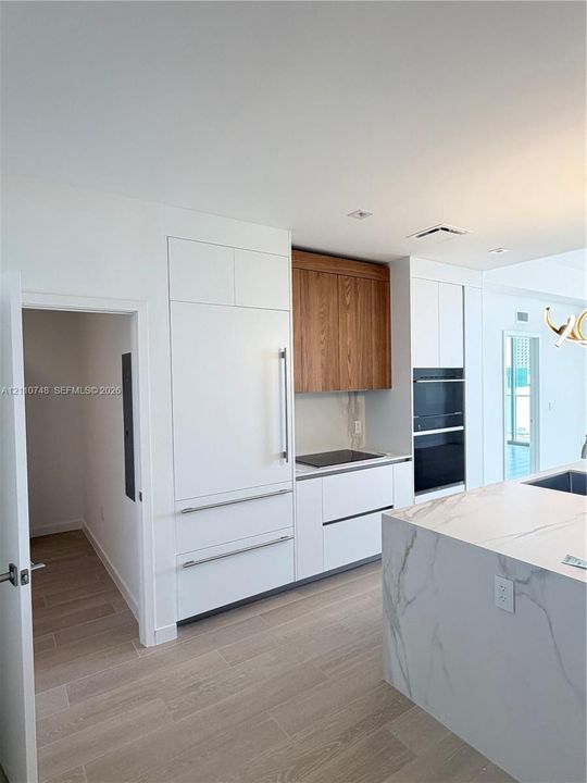 Kitchen featuring a stone-surface island, built-in integrated refrigerator, wall-mounted double oven, custom flat-panel cabinetry with wood-finish upper accents, and light-toned wood-finish flooring