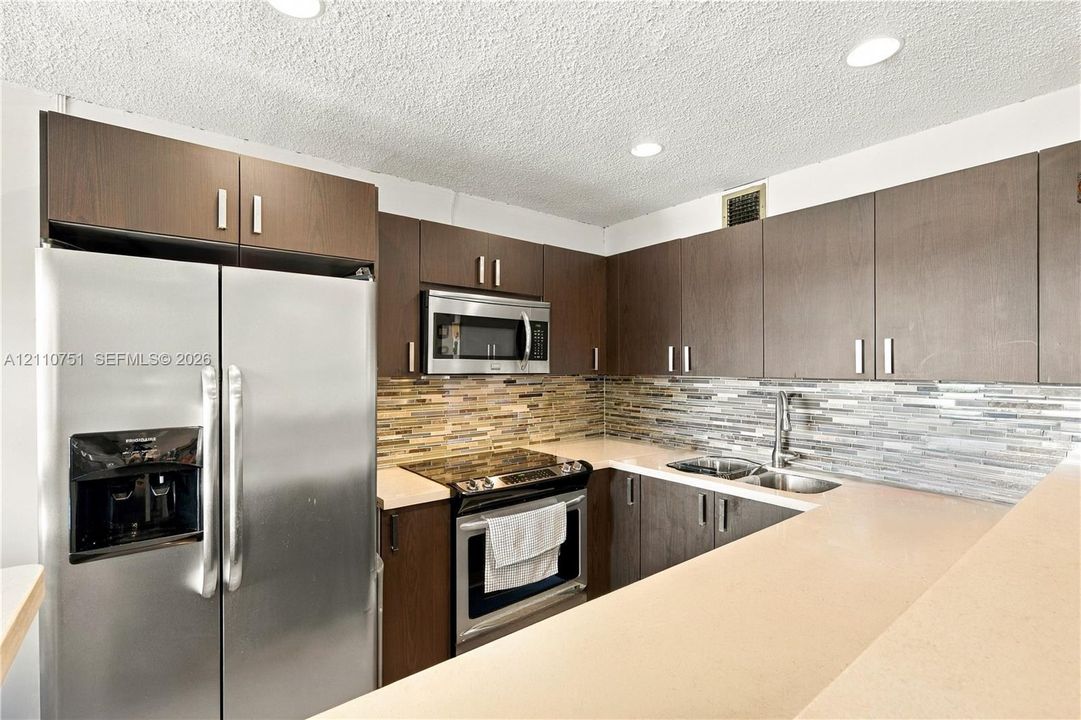 Kitchen featuring flat-panel wood-finish cabinetry, stainless steel appliances, multi-tonal linear tile backsplash, solid-surface countertops, and recessed lighting